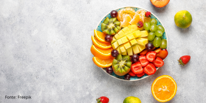 Tigela com diversas frutas cortadas, morangos, kiwis, laranjas, uvas e mangas, com frutas colocadas em meia-lua fora do pote em um fundo cinza, cimento queimado