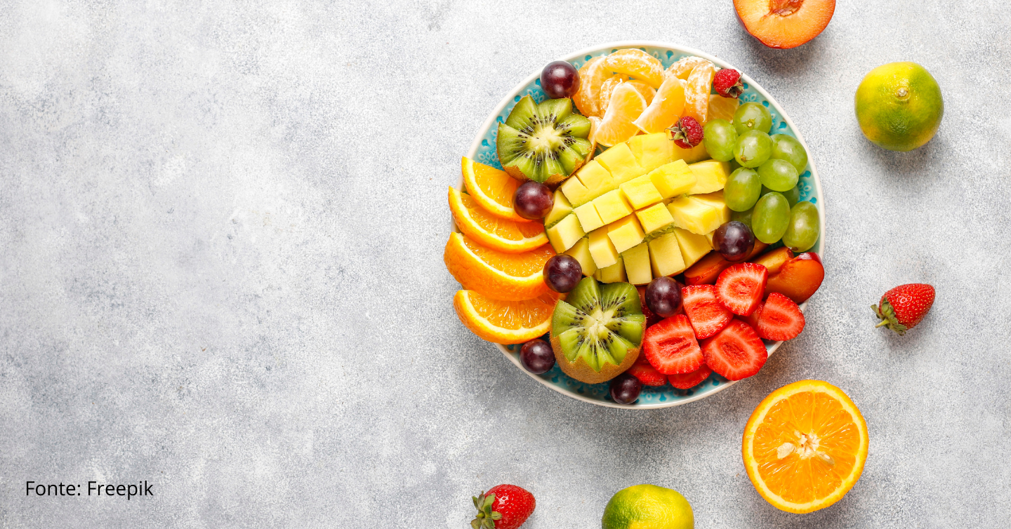 Tigela com diversas frutas cortadas, morangos, kiwis, laranjas, uvas e mangas, com frutas colocadas em meia-lua fora do pote em um fundo cinza, cimento queimado